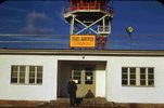 A pilot standing in front of the entrance to a building at Tegel Airfield. The red tower on the building can be seen in the background. The colour photograph also shows an orange sign with the words “Tegel Airfield” on it above the entrance.