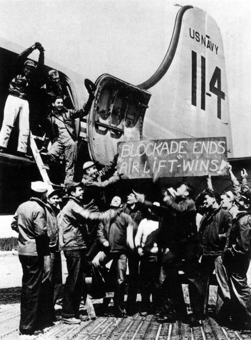 A group of men hold a sign reading, “Blockade Ends Airlift Wins” standing near the cargo hold of a plane.