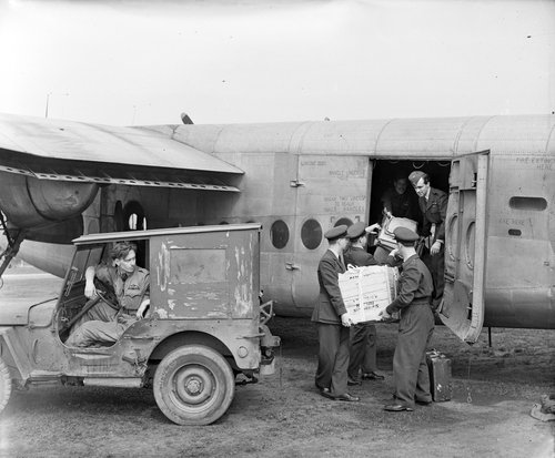 Men loading crates into an aeroplane. A jeep parked nearby.