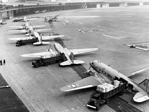 Reihe von zweimotorigen Transportflugzeugen, die in einer Reihe auf einem Flugplatz geparkt sind, wobei jeweils ein Lastwagen zum Verladen von Vorräten geparkt ist.