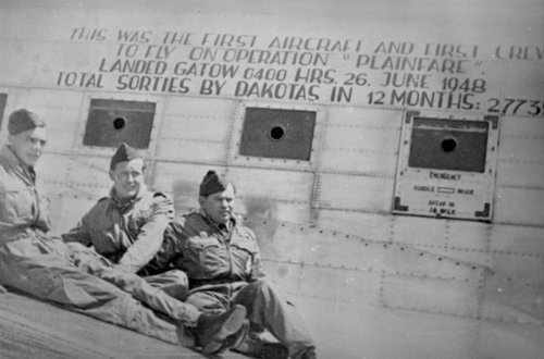 Three men in RAF uniform on the wing of a transport aeroplane.  Painted on the aeroplane is: This was the first aircraft and first crew to fly on Operation “Plainfare”.  Landed Gatow 0400 hrs 26 June 1948.  Total sorties by Dakotas in 12 months: 27735.