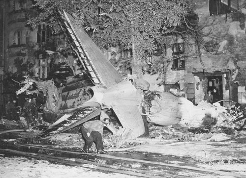 Wrecked tail of an aeroplane seen at an angle with tree and building in the background.