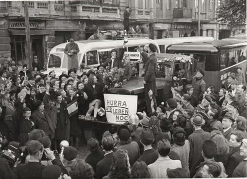 Rund 100 Personen umringen einen LKW, an dem Blumen und das Plakat mit der Aufschrift „Hurra wir leben noch“ befestigt ist, 12. Mai 1949.