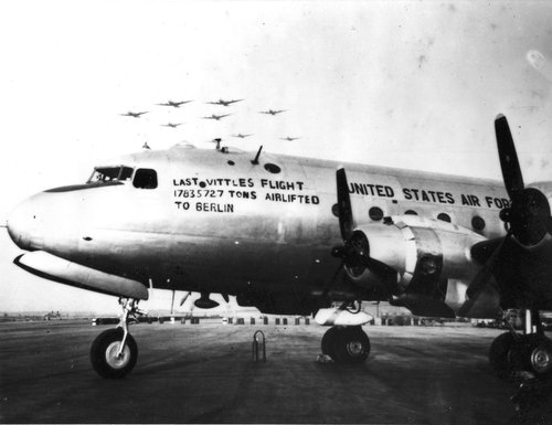 Ein viermotoriges Frachtflugzeug steht auf einer Start- und Landebahn, während eine Formation von Transportflugzeugen darüber fliegt. Auf dem Flugzeug steht die Aufschrift „Letzter Vittles Flug. 17.835.727 Tonnen nach Berlin geflogen.“