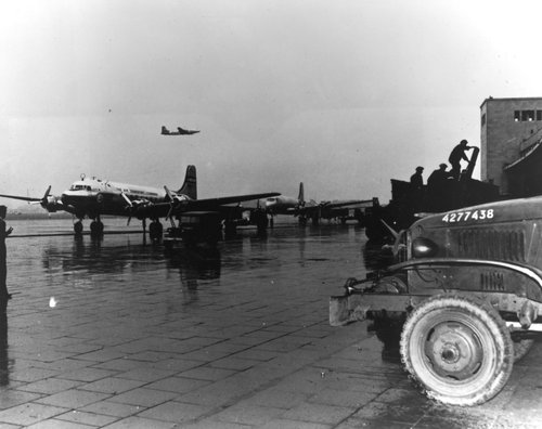 A panoramic photo of an aircraft on the ground, a plane in the air, and the front a ground vehicle on a runway.