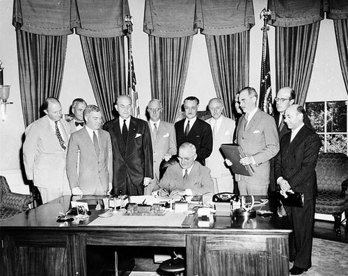 A man sits writing at a desk, with a number of other men standing behind him.