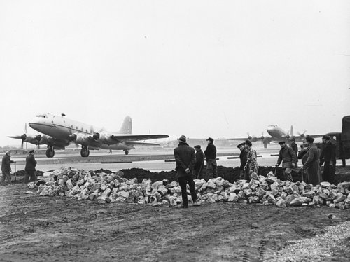 A group of people in a shallow hole on an airfield, watching as two four-engined transport aeroplanes taxiing past in the background.
