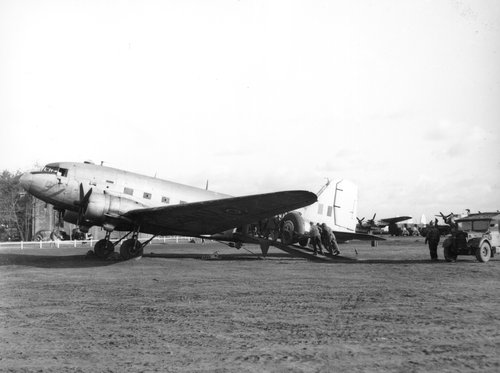 View of a twin-engined transport aeroplane on the ground, with men pushing a vehicle up a ramp to the aircraft’s rear side door.